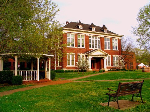 A historic red brick building with a gazebo and bench in a green park setting, surrounded by trees and shrubs.