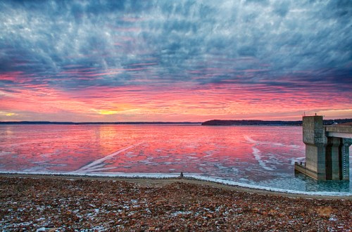 A vibrant sunset over a frozen lake, with colorful clouds reflecting on the icy water and a pier in the foreground.
