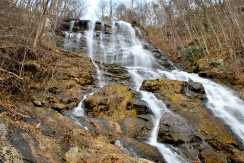 A cascading waterfall flows down a rocky cliff, surrounded by bare trees and mossy stones.