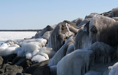 Ice-covered rocks along a shoreline, with frozen formations glistening in the sunlight against a clear blue sky.