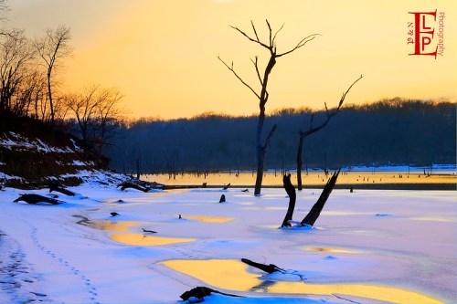 A serene winter landscape with a frozen lake, snow-covered shore, and bare trees against a colorful sunset sky.