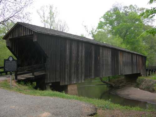 A wooden covered bridge spans a calm creek, surrounded by lush green trees and a gravel path.