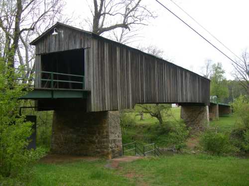 A wooden covered bridge spans a small creek, supported by stone pillars, surrounded by greenery and trees.