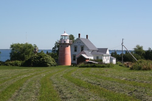 A pink lighthouse beside a white house, surrounded by green fields and a blue lake under a clear sky.
