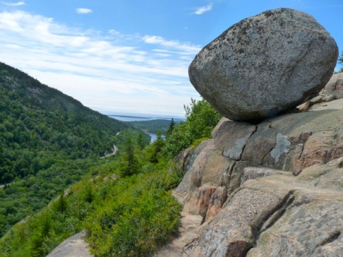 A large boulder perched on rocky terrain, overlooking a lush green valley and a winding river under a blue sky.
