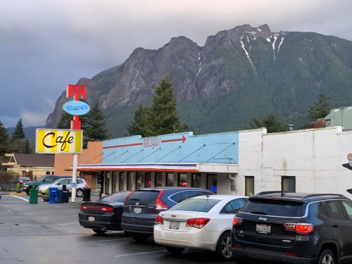 A colorful cafe building with a mountain backdrop and parked cars in the foreground.