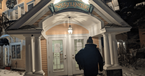 A person walks towards a warmly lit entrance of a building labeled "Nestlenook" in a snowy setting at night.