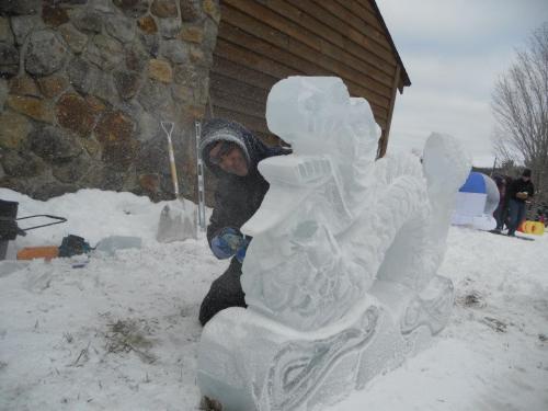 A person carves a detailed ice sculpture in a snowy outdoor setting, with a stone wall and wooden structure in the background.