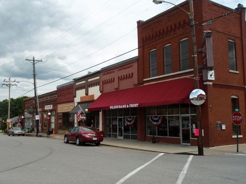 A small town street featuring brick buildings, a bank with an awning, and American flags, under a cloudy sky.