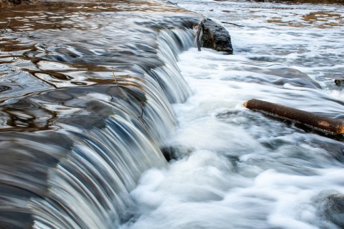 A gentle waterfall cascades over rocks, creating rippling water and reflections in a serene natural setting.