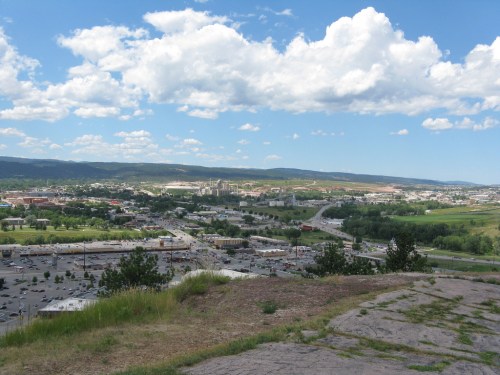 A panoramic view of a cityscape with green fields, roads, and mountains under a blue sky with fluffy clouds.