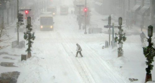 A person walks through a snowy street with low visibility, surrounded by snow-covered buildings and traffic lights.