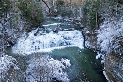 A serene winter scene of a waterfall cascading over rocks, surrounded by snow-covered trees and a tranquil river.