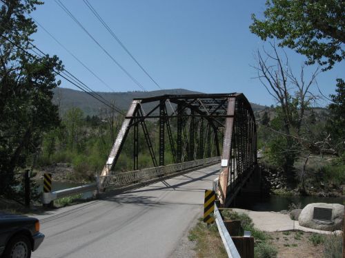 A metal arch bridge spans a river, surrounded by trees and mountains under a clear blue sky.