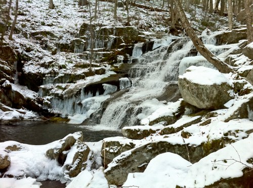 A serene winter scene of a cascading waterfall surrounded by snow-covered rocks and trees.