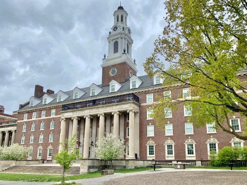 Historic brick building with a clock tower, large columns, and trees in bloom under a cloudy sky.