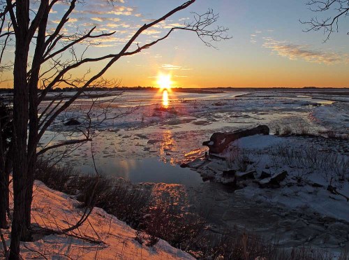 A serene winter sunset over a frozen river, with silhouettes of trees and ice reflecting the warm colors of the sky.