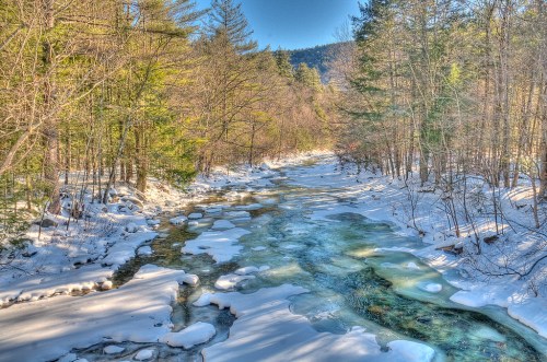 A serene winter landscape featuring a partially frozen river surrounded by snow-covered trees and mountains in the background.