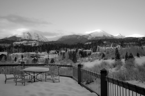 A snowy landscape with mountains in the background, featuring a table and chairs on a deck. Black and white photo.