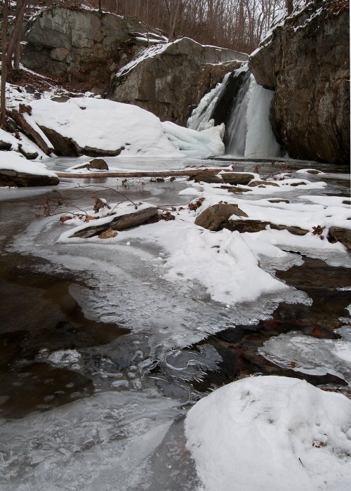 A snowy landscape featuring a frozen waterfall and icy stream surrounded by rocky terrain and bare trees.