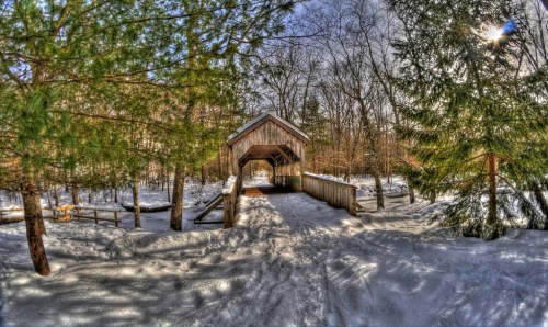 A snow-covered path leads to a wooden covered bridge surrounded by trees in a winter landscape.
