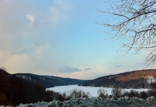 A serene winter landscape featuring a frozen lake surrounded by snow-covered hills and a clear blue sky.