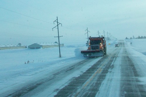 A snowplow clears a snowy road lined with power poles and buildings under a pale winter sky.