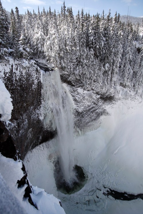 A snowy waterfall cascades down rocky cliffs, surrounded by tall evergreen trees in a winter landscape.
