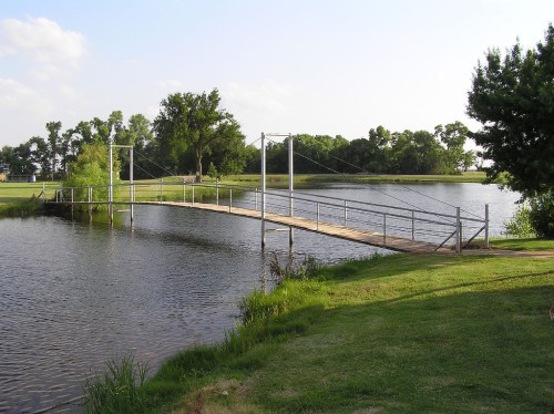 A wooden suspension bridge spans a calm lake, surrounded by greenery and trees under a clear sky.