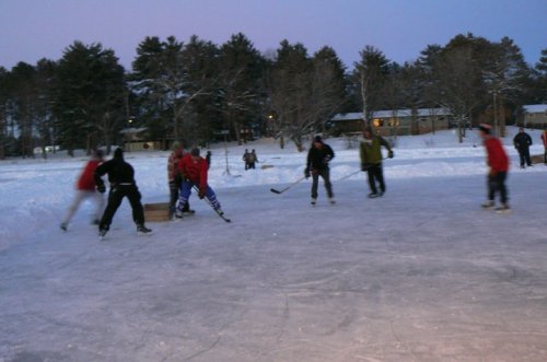 A group of people playing ice hockey on a snowy outdoor rink at dusk, surrounded by trees and a few spectators.