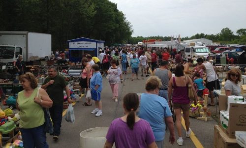 A bustling outdoor flea market with many people browsing various stalls and items along a crowded pathway.