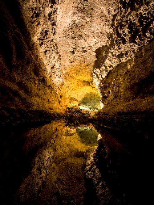 A dimly lit cave with a reflective water surface and textured rock walls, leading to a bright opening in the distance.