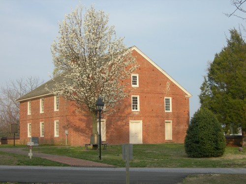A brick building with a flowering tree in front, surrounded by grass and a pathway, under a clear sky.
