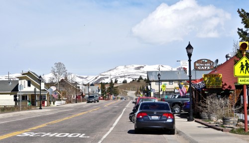 A small town street with parked cars, shops, and snow-capped mountains in the background under a partly cloudy sky.