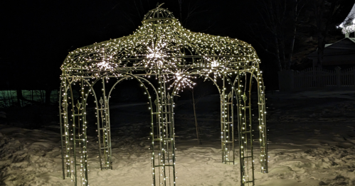 A decorative gazebo covered in twinkling white lights, set against a dark winter night. Snow is visible on the ground.
