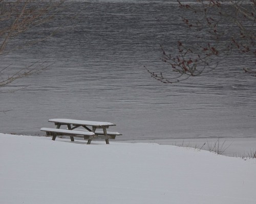 A snowy landscape featuring a picnic table by a calm, icy lake.