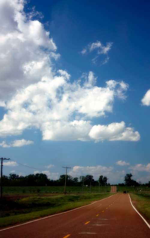 A winding road stretches into the distance under a blue sky filled with fluffy white clouds. Green fields line the sides.