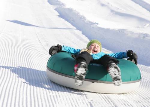 A child joyfully sledding down a snowy hill on a green inner tube, wearing a bright blue jacket and a green hat.