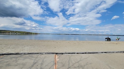 A serene beach scene with calm water, sandy shore, and a few people enjoying the outdoors under a partly cloudy sky.