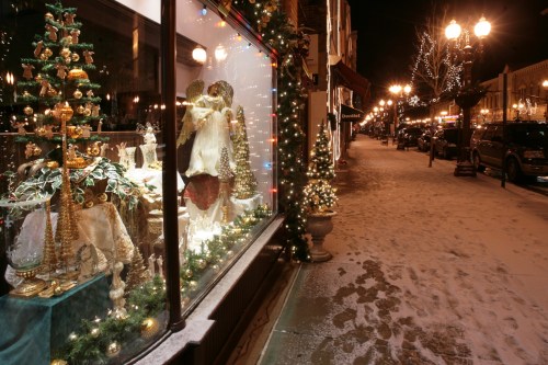 A festive street scene at night, featuring a decorated shop window and snow-covered sidewalks with holiday lights.