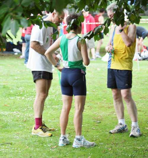 Three runners in athletic gear converse in a grassy area, surrounded by trees and a crowd in the background.