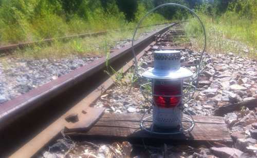 A vintage lantern sits on a wooden plank beside overgrown railway tracks, surrounded by grass and gravel.