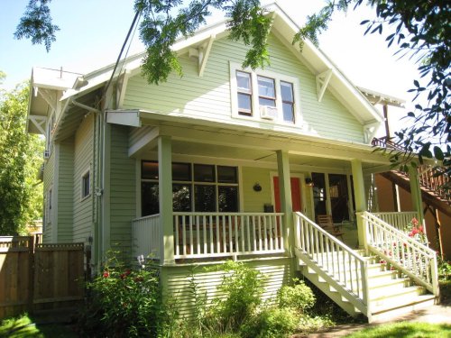 A light green house with a porch, surrounded by greenery and flowers, under a clear blue sky.