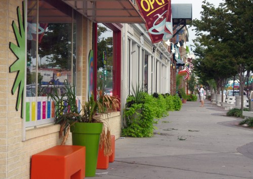 A colorful sidewalk scene with potted plants, storefronts, and a person walking in the distance on a cloudy day.