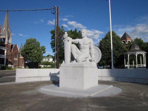 A white stone statue of a seated figure holding a sword, surrounded by trees and buildings under a blue sky.