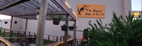 Exterior view of The Roost Bar & Grill, featuring a wooden patio with hanging plants and a large sign.