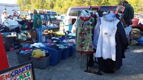 A bustling outdoor market with clothing racks, bins of clothes, and shoppers browsing among various items.