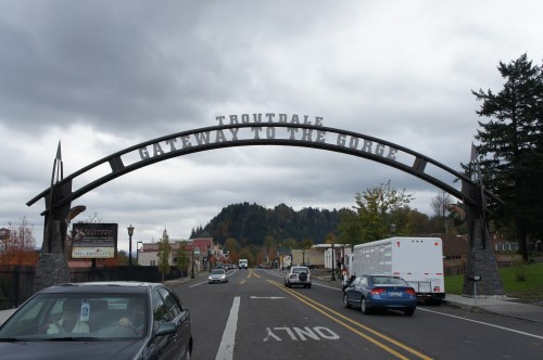 Archway sign reading "Troutdale Gateway to the Gorge" over a street with cars and trees under a cloudy sky.