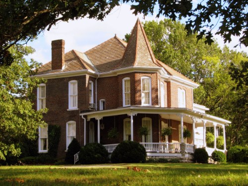 A charming brick house with a pointed roof, surrounded by greenery and featuring a spacious front porch.