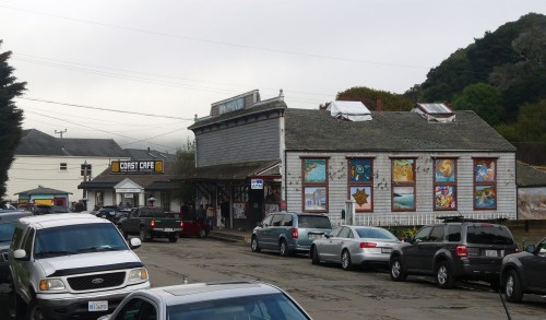 A street view of a quaint building with colorful murals, surrounded by parked cars on a cloudy day.